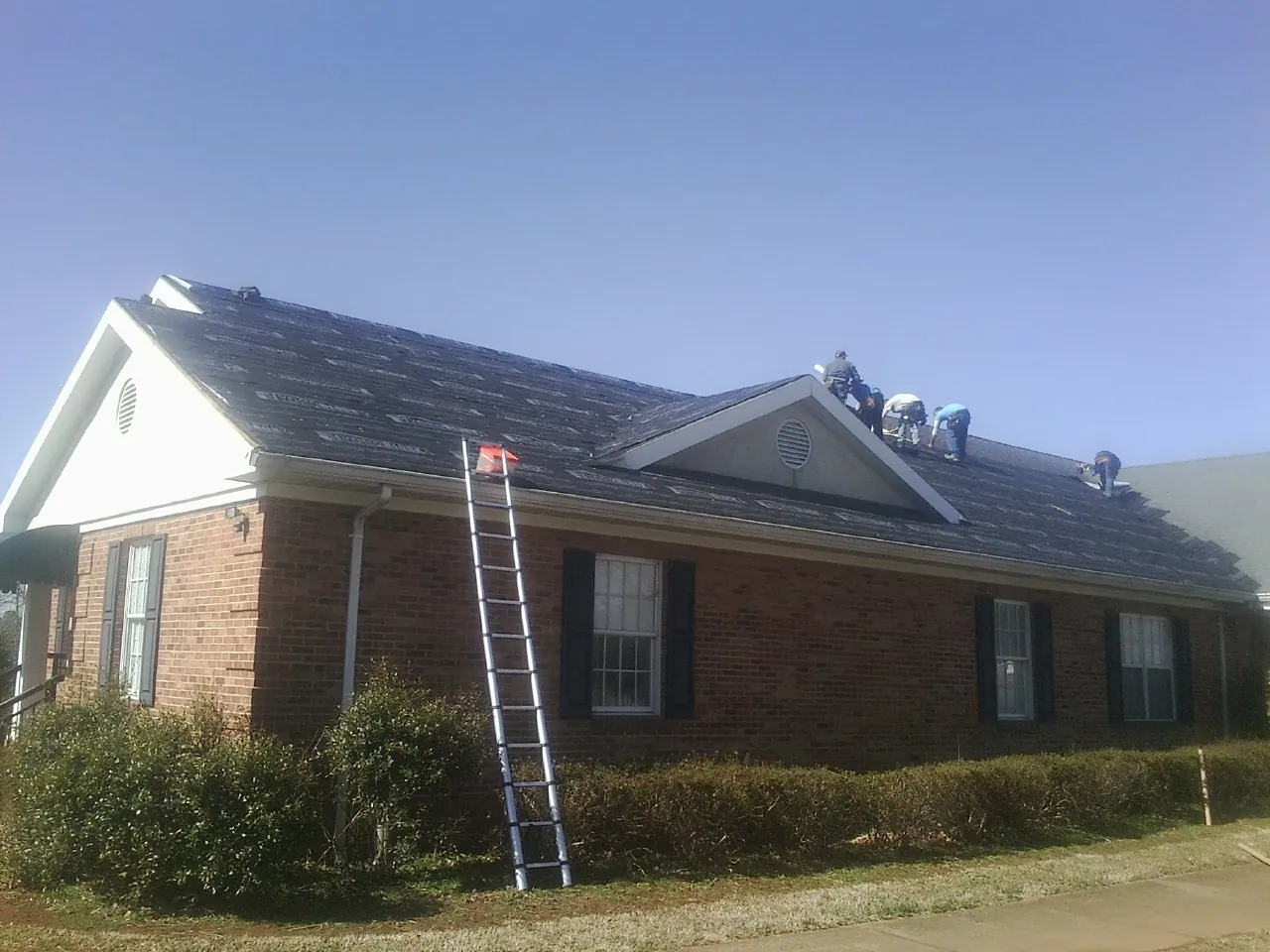 Crew tearing shingles off a brick ranch roof with a ladder set — Emergency Roof Repair work by our crew in Lennon