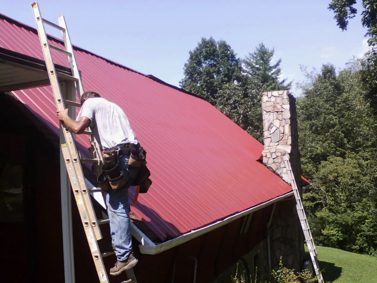 Roofer on a ladder beside a red metal roof and stone chimney — Soffit & Fascia Repair by Roofing Brothers in Lennon